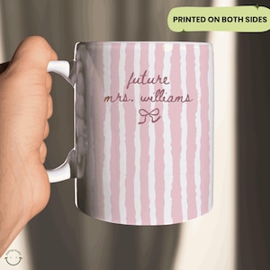 May include: White ceramic mug with pink vertical stripes. The mug has the text "future mrs. williams" in a script font, with a bow graphic. The words "printed on both sides" are in a green rectangle.