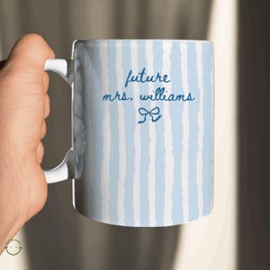 May include: White ceramic mug with a blue and white striped pattern. The mug features the text "future mrs. williams" in a handwritten font, along with a bow graphic. The mug has a white handle.