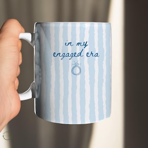 May include: White ceramic mug with a light blue and white striped pattern. The mug has the text "in my engaged era" and a diamond ring graphic in blue. The mug is held by a hand.