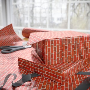 May include: Two gift boxes wrapped in red brick-patterned paper are stacked on a table. A roll of matching wrapping paper and a pair of scissors are visible. A black ribbon is draped across the table, adding a decorative touch to the scene.