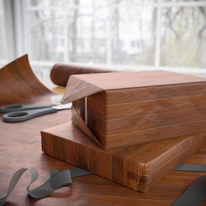 May include: Two rectangular boxes wrapped in brown wood grain patterned paper. The paper features a realistic dark brown wood texture with vertical grain lines. A pair of grey scissors and a loose grey ribbon are visible near the wrapped boxes.