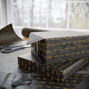 Pode incluir: Dois presentes embrulhados com papel preto e padrões geométricos dourados. Um rolo de papel de embrulho a condizer, tesouras e uma fita preta estão sobre a mesa. Os presentes estão empilhados, um parcialmente desembrulhado.