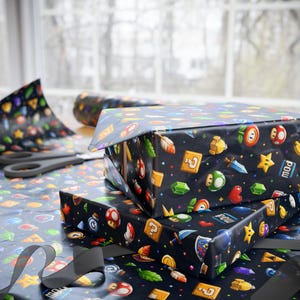 May include: Two wrapped gifts with black wrapping paper featuring colorful video game icons. The gifts are stacked on top of each other. Scissors and a roll of matching wrapping paper are visible in the background. A black ribbon is in the foreground.