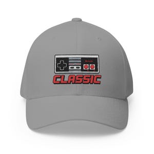 May include: A gray baseball cap featuring a retro video game controller design. The controller is black, red, and white, with the word "CLASSIC" in red block letters below. A stylish accessory for gamers.