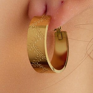May include: A gold-toned hoop earring featuring a flat, wide band with an engraved pattern of delicate, repeating leaves. The earring has a polished metallic finish and a hinged closure mechanism, worn through the earlobe in a circular shape.