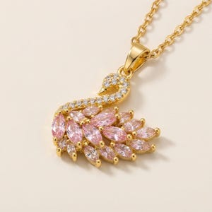 Può includere: Una collana con pendente a forma di cigno color oro. Il corpo del cigno è adornato con pietre preziose rosa e bianche, e il collo è incastonato con cristalli trasparenti. La catena della collana è anch'essa color oro.