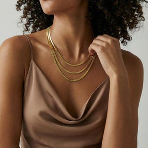 Op de afbeelding: Een gelaagde goudkleurige ketting met drie strengen van verschillende lengtes. De ketting heeft een plat slangenkettingontwerp. Het model draagt een bruine satijnen camisole.