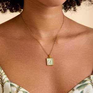 Può includere: Collana dorata con un ciondolo quadrato. Il ciondolo presenta uno sfondo verde chiaro con un disegno floreale bianco e rosa. La collana è indossata intorno al collo.