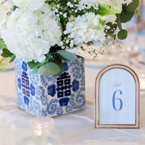 May include: A display of white flowers, such as roses and hydrangeas, in a blue and white patterned vase. A gold-framed table number 6 is visible, indicating a wedding or event. The vase has a Chinese design.