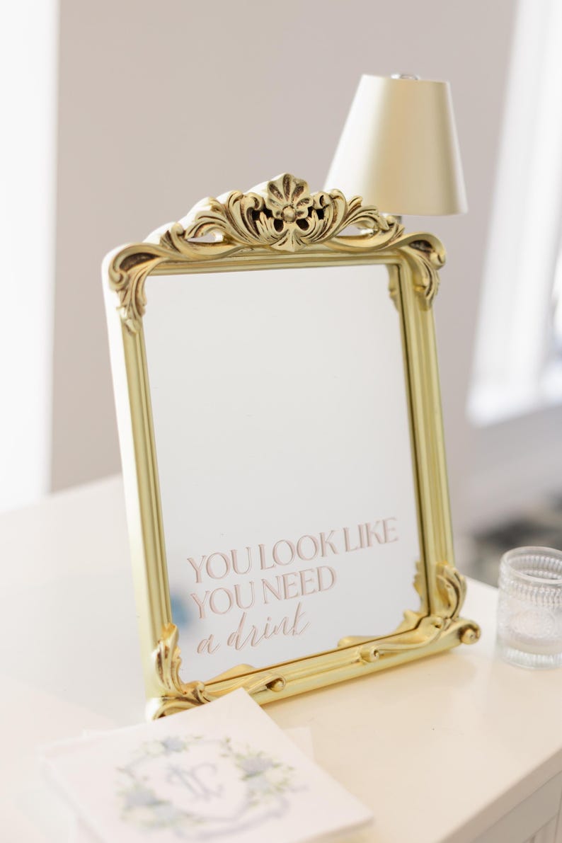 Puede incluir: Espejo con marco dorado y detalles ornamentales. El espejo refleja el texto "YOU LOOK LIKE YOU NEED a drink" en escritura cursiva. Se ven una l&aacute;mpara peque&ntilde;a y un vaso decorativo.