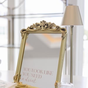 Puede incluir: Espejo dorado con marco ornamentado, mostrando el texto rosa "YOU LOOK LIKE YOU NEED a drink". Una peque&ntilde;a l&aacute;mpara y un vaso est&aacute;n al lado, sugiriendo un ambiente de bar o evento.