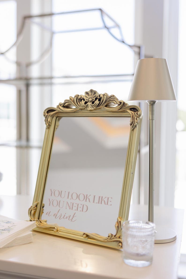 Puede incluir: Espejo dorado con marco ornamentado, mostrando el texto rosa "YOU LOOK LIKE YOU NEED a drink". Una peque&ntilde;a l&aacute;mpara y un vaso est&aacute;n al lado, sugiriendo un ambiente de bar o evento.