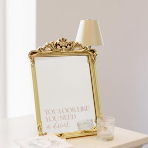 Puede incluir: Un espejo dorado con el texto "YOU LOOK LIKE YOU NEED a drink" se muestra sobre una superficie blanca. Tambi&eacute;n hay una peque&ntilde;a l&aacute;mpara y un vaso decorativo. El espejo tiene detalles ornamentados, la est&eacute;tica es elegante.