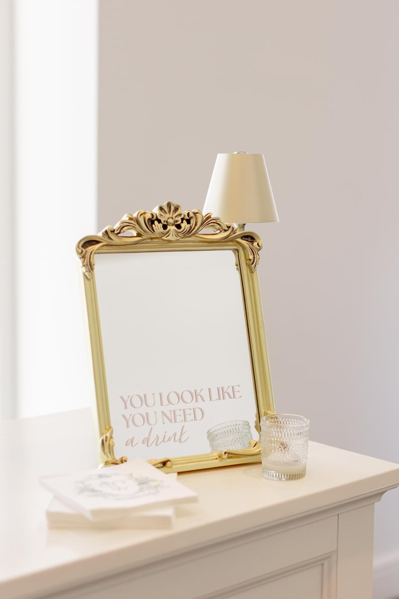 Puede incluir: Un espejo dorado con el texto "YOU LOOK LIKE YOU NEED a drink" se muestra sobre una superficie blanca. Tambi&eacute;n hay una peque&ntilde;a l&aacute;mpara y un vaso decorativo. El espejo tiene detalles ornamentados, la est&eacute;tica es elegante.