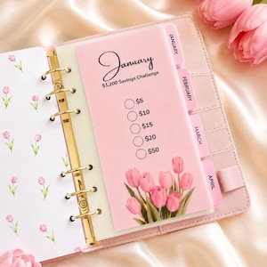 May include: A pink personal planner with gold rings featuring a January $1,200 Savings Challenge insert. The page displays five circular checkboxes labelled $5, $10, $15, $20, and $50, decorated with a bouquet of pink tulips at the bottom.
