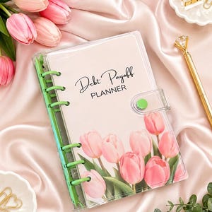 May include: A clear plastic debt payoff planner with a light pink cover featuring a printed design of pink tulips. The planner has bright green binder rings and a green snap closure. The text Debt Payoff PLANNER is printed in black ink.