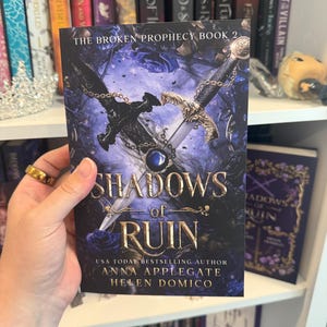 May include: A book cover for "Shadows of Ruin" features a dark, fantasy-themed design. The title is in gold lettering, with a sword and chain design. The author's names are also visible. The book is part of "The Broken Prophecy" series.
