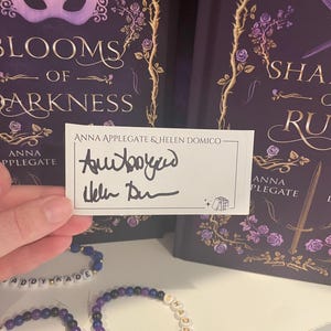 Puede incluir: Una tarjeta firmada sostenida frente a un libro titulado "Blooms of Darkness" y una pulsera. La tarjeta está firmada por Anna Applegate y Helen Domico. La pulsera tiene cuentas de letras blancas que deletrean "Shadow of Ruin".