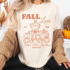 May include: An ivory t-shirt featuring a burnt orange graphic with the text FALL in Love, est. 2026, Future Mrs. Willson, and Boston, MA. The design includes a champagne coupe, pumpkins, an acorn, and an open ring box with a ring inside.