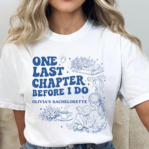 May include: White t-shirt with a neon blue design. The shirt features the text "ONE LAST CHAPTER BEFORE I DO" and "OLIVIA'S BACHELORETTE" with illustrations of a ring, flowers, books, and a woman reading.