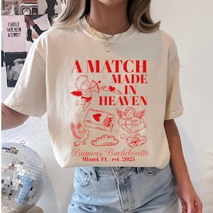 以下が含まれることがあります： クリーム色のTシャツに赤い文字とイラスト。テキストは「A Match Made in Heaven」で、キューピッドとハートのデザイン。下には「Bianca's Bachelorette, Miami, FL est. 2025」と書かれています。