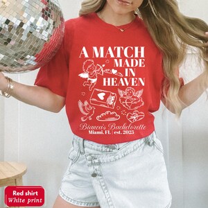 以下が含まれることがあります： 「A MATCH MADE IN HEAVEN」と書かれた白いグラフィックの赤いTシャツ。デザインには、天使、ハート、そして「Bianca's Bachelorette Miami, FL est. 2025」という文字が含まれています。