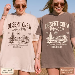May include: Two women wearing ivory and espresso-colored t-shirts with a desert-themed graphic. The shirts feature the text "DESERT CREW Before I Do" with a cactus, sun, and mountain design, and "Ashley's Bachelorette, Charleston, AZ."