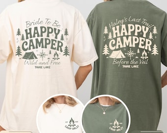 May include: Two oversized t-shirts, one cream and one khaki green, display camping-themed designs. The cream shirt reads "Bride To Be HAPPY CAMPER Wild and Free TAHOE LAKE." The khaki green shirt says "Haley's Last Trail HAPPY CAMPER Before the Veil TAHOE LAKE."