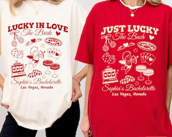 May include: Two oversized t-shirts, one ivory and one red, with cherry-colored graphics. The ivory shirt reads "LUCKY IN LOVE The Bride" and the red shirt says "JUST LUCKY The Bach". Both shirts feature casino-themed illustrations and text: "Sophia's Bachelorette Las Vegas, Nevada."