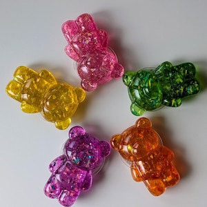May include: Five colorful, glitter-filled gummy bear-shaped phone grips. The bears are in shades of pink, yellow, green, purple, and orange. Each grip has a clear, circular adhesive base. The bears are arranged in a circular pattern against a white background.
