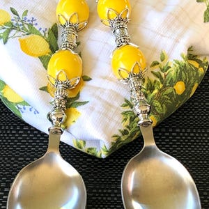 Puede incluir: Un par de cucharas de servir con mangos de cuentas amarillas y cabezas de cuchara plateadas. Las cucharas descansan sobre un paño blanco con un estampado de limones y flores. Los mangos tienen detalles decorativos plateados.