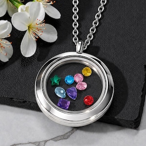 Può includere: Una collana con un medaglione d'argento e un centro trasparente pieno di pietre preziose colorate. Le pietre preziose includono una stella verde, gemme blu, rosa, gialle e rosse. La collana è su una superficie nera con fiori bianchi.
