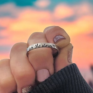 May include: A silver ring with the inscription "I am the storm" and a small heart symbol. The ring is worn on a person's finger.