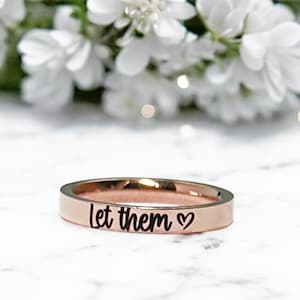 May include: A rose gold ring with the words "let them" and a small heart engraved on the band.