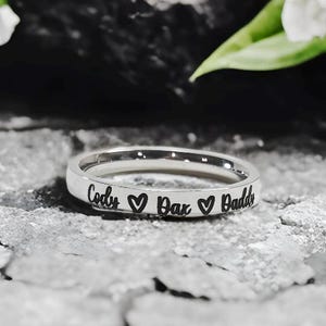 May include: A silver ring with the names "Cody", "Dax", and "Daddi" engraved, separated by heart symbols. The ring is set against a textured gray surface, with white flowers in the background.