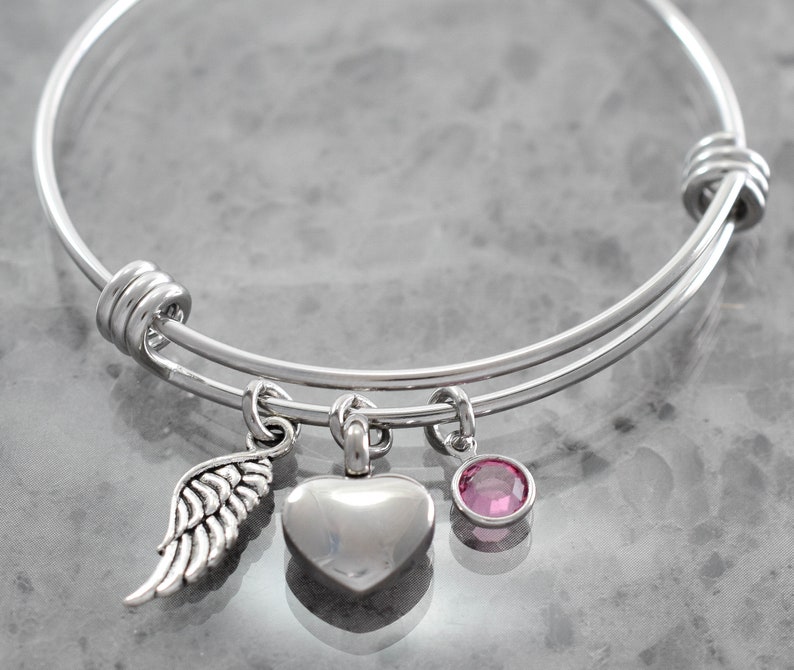 Puede incluir: Un brazalete expandible de plata con tres dijes: una urna de cremaci&oacute;n en forma de coraz&oacute;n, un ala de &aacute;ngel peque&ntilde;a y un cristal rosa.