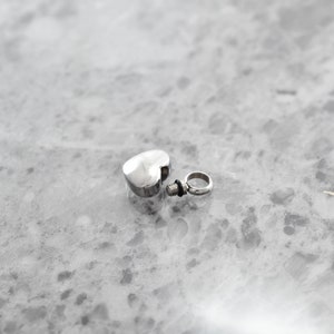 Puede incluir: Un colgante de cremaci&oacute;n de plata en forma de coraz&oacute;n con una tapa de rosca y un peque&ntilde;o anillo para sujetarlo a una cadena.