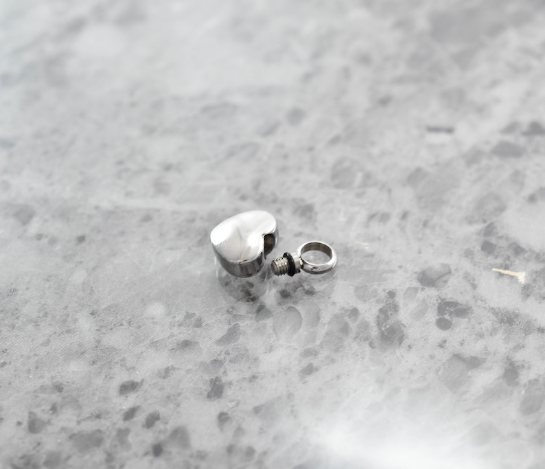 Puede incluir: Un colgante de cremaci&oacute;n de plata en forma de coraz&oacute;n con una tapa de rosca y un peque&ntilde;o anillo para sujetarlo a una cadena.