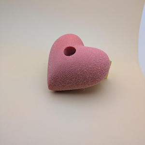 May include: A textured, heart-shaped candle holder in a dusty rose color. The candle holder has a small hole in the center for a candle. The surface has a speckled appearance. The item is on a light beige background.