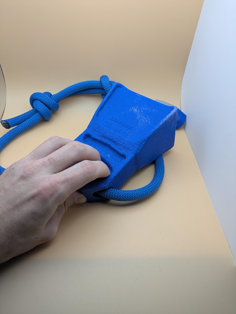 Puede incluir: Un agarre de escalada azul con una cuerda adjunta. El agarre tiene forma rectangular con el texto "Cowboy Climbing" impreso en el lateral. La cuerda es de color azul a juego y est&aacute; anudada en un extremo.