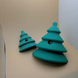 May include: Two teal-colored Christmas tree-shaped objects. The trees have a textured surface and a small, round detail at the top. One tree is larger than the other. The trees are on a light tan background.