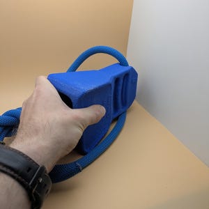 Puede incluir: Un juguete azul para perros con un asa de cuerda. El juguete tiene una forma rectangular s&oacute;lida con un asa curva hecha de cuerda azul. El juguete est&aacute; siendo sostenido por una persona.