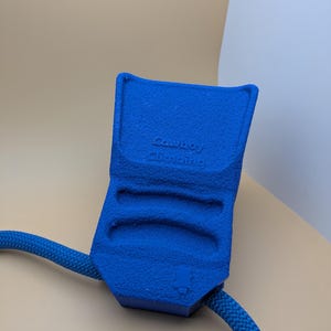 Puede incluir: Un dispositivo de escalada azul texturizado con el texto "Cowboy Climbing" est&aacute; conectado a una cuerda azul. El dispositivo tiene tres ranuras para los dedos y un peque&ntilde;o logotipo. El fondo es beige y blanco neutro.