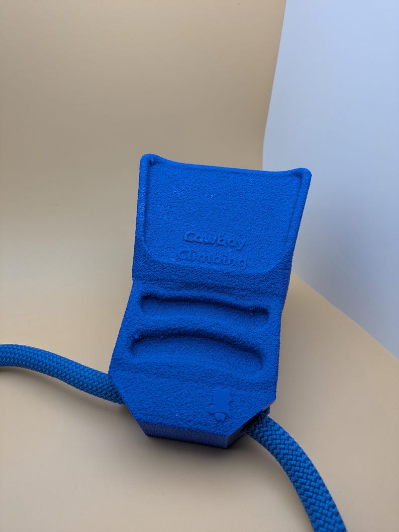 Puede incluir: Un dispositivo de escalada azul texturizado con el texto "Cowboy Climbing" est&aacute; conectado a una cuerda azul. El dispositivo tiene tres ranuras para los dedos y un peque&ntilde;o logotipo. El fondo es beige y blanco neutro.