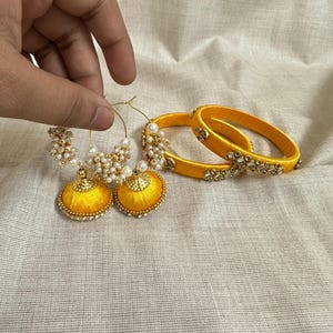 Puede incluir: Un par de pendientes amarillos de hilo de seda con detalles de perlas y pulseras amarillas a juego. Los pendientes tienen forma de campana con una parte superior dorada y un aro adornado con perlas. Las pulseras están adornadas con pequeños elementos decorativos.