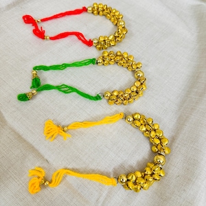 May include: Three decorative bell bracelets with red, green, and yellow cords. Each bracelet features a series of small, gold-colored bells. The bracelets are arranged on a white fabric background.