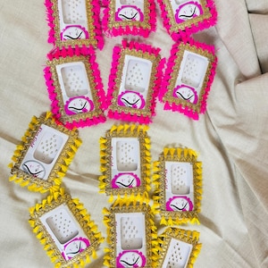 May include: A collection of decorative phone cases with a rectangular design. The cases feature a white interior, a gold border, and are adorned with pink or yellow tassels. Each case has a small, circular design with a face.