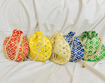 Può includere: Cinque sacchetti con coulisse colorati in rosso, giallo, oro, blu e verde. Ogni sacchetto presenta un motivo geometrico con dettagli dorati ed è adornato con un bordo e un cordoncino di perle. I sacchetti sono disposti su un tessuto bianco.