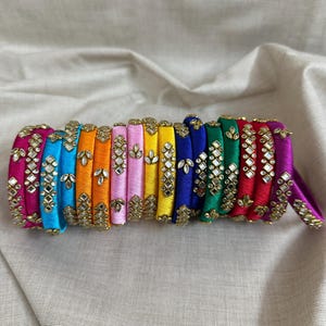 May include: A collection of colorful bangles in a variety of hues, including pink, blue, orange, yellow, and green. Each bangle is adorned with gold-colored embellishments and small mirrored accents, creating a decorative and eye-catching design.