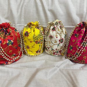 Puede incluir: Cuatro bolsas de cordón coloridas. Las bolsas son rojas, amarillas, blancas y rosas, cada una adornada con bordados florales y un ribete de cuentas de perlas. Los cordones son dorados. Las bolsas están dispuestas sobre un fondo de tela blanca.